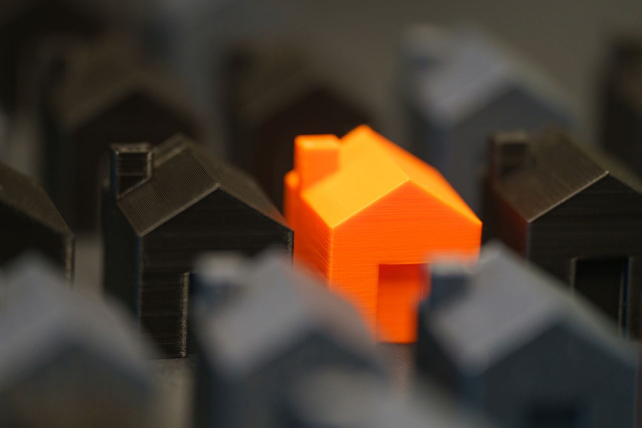 A vibrant orange model house stands out among black miniature houses, symbolizing real estate concepts.
